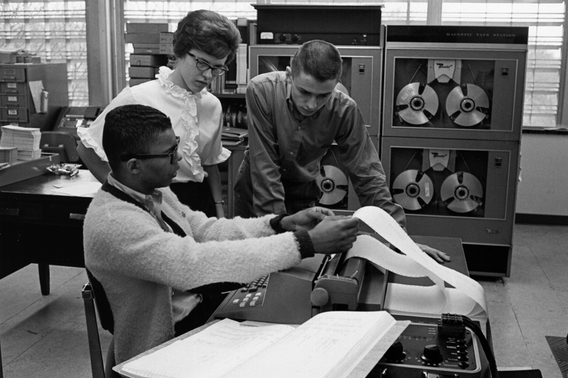 Three students look at a paper emerging from a typewriter.